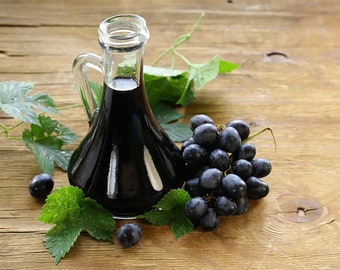 May include: A clear glass bottle filled with dark red balsamic vinegar, surrounded by green grape leaves and a bunch of dark purple grapes on a wooden surface.