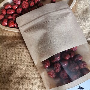 Puede incluir: Una bolsa de papel marr&oacute;n con una ventana de pl&aacute;stico transparente llena de d&aacute;tiles rojos secos. La bolsa tiene una etiqueta blanca con el texto "All Native Natural Foods" y un peque&ntilde;o logotipo de una planta.