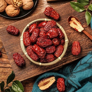 May include: A close-up of a bowl filled with dried red dates, a traditional Chinese ingredient. The dates are arranged in a circular pattern and are surrounded by green leaves and walnuts.