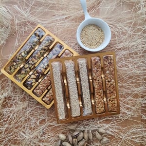 Op de afbeelding: Twee gouden bakken met verschillende sesam- en notenrepen. De bakken zijn gevuld met rijen repen, elk met een andere combinatie van sesamzaad, noten en honing. De bakken staan op een bruin oppervlak met een witte kom met sesamzaad en een stapel pistachenoten.