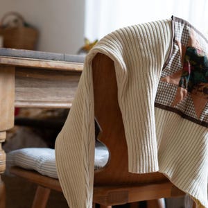 Puede incluir: Un suéter de punto acanalado color crema drapeado sobre una silla de madera. La silla tiene respaldo decorativo y está junto a una mesa. Una colcha de retazos con telas marrones y beige está sobre la silla. La escena está iluminada por luz natural.