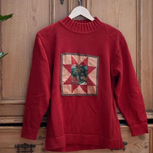 Op de afbeelding: Een rode trui met een hoge halslijn en lange mouwen. De trui heeft een vierkante patch met een sterontwerp in rood, beige en groen. De trui hangt aan een houten hanger.