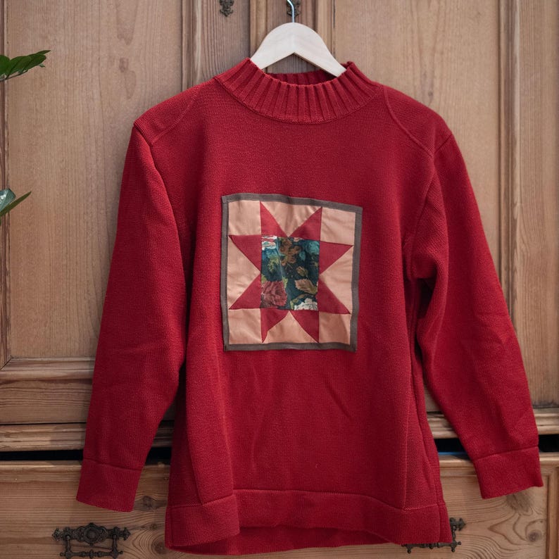 Op de afbeelding: Een rode trui met een hoge halslijn en lange mouwen. De trui heeft een vierkante patch met een sterontwerp in rood, beige en groen. De trui hangt aan een houten hanger.