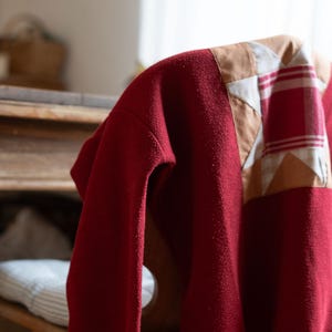 Puede incluir: Un suéter rojo oscuro con un diseño de patchwork en el hombro. El patchwork presenta tela a cuadros beige, blanca y roja. El suéter está drapeado sobre una silla de madera, mostrando su textura y color.