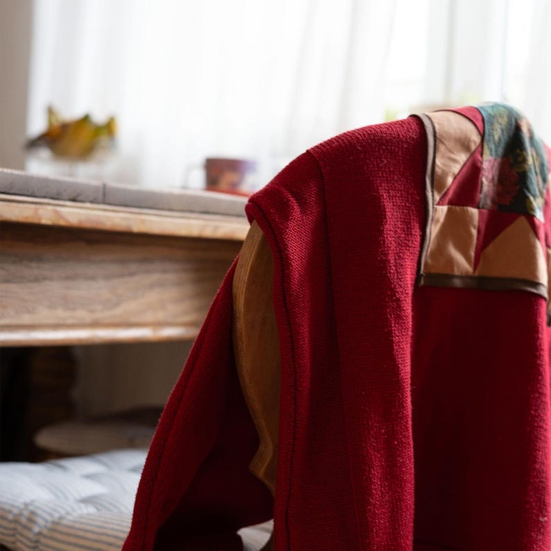 Op de afbeelding: Een rode, getextureerde jas met een patchwork-ontwerp op de schouder hangt over een houten stoel. Het patchwork heeft beige, bruine en bloemmotiefstof. De stoel heeft een lichtbruin houten frame en een gestreept kussen.