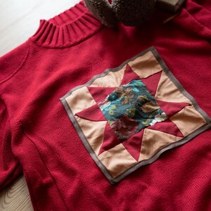 Peut inclure: Un pull rouge avec un écusson décoratif en forme d'étoile. L'écusson présente un carré central avec un motif floral dans les tons de bleu, vert et rouge. Les pointes de l'étoile sont rouges et beiges, bordées d'un liseré marron. Un cache-oreilles marron est en arrière-plan.