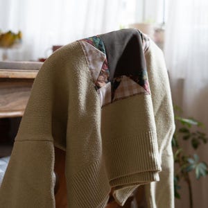 Könnte beinhalten: Ein hellgrüner Pullover mit einem Patchwork-Design auf den Schultern, drapiert über einem Holzstuhl. Der Patchwork-Stoff weist Blumen- und Karomuster auf und verleiht dem Strick einen Vintage-Touch.