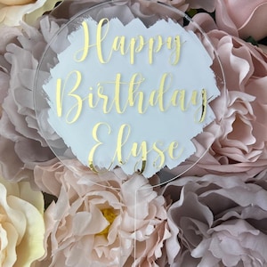 May include: A clear acrylic cake topper with a white background and gold lettering. The topper says "Happy Birthday Elyse" with a brushstroke design.