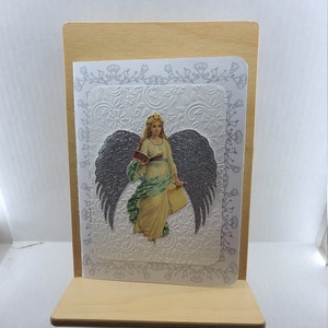 May include: A white card with a silver embossed border and a floral design. The card features a vintage image of an angel with wings holding a book. The card is displayed on a wooden easel.