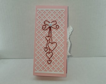 May include: A pink card with a white lattice design and a red glitter bow with a pearl in the center. Hanging from the bow are five red glitter hearts.