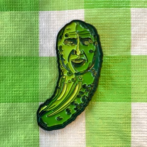 Puede incluir: Un pin de esmalte verde con forma de pepinillo con una cara. El pepinillo tiene una expresión sonriente y está sobre un fondo a cuadros verdes y blancos.