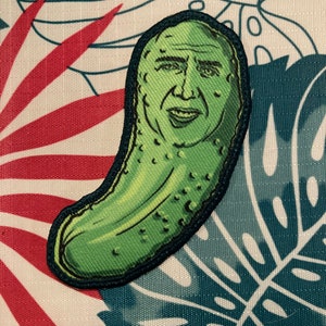 Puede incluir: Un parche bordado verde de un pepinillo con la cara de un hombre. El pepinillo tiene la forma de un pepinillo encurtido tradicional.