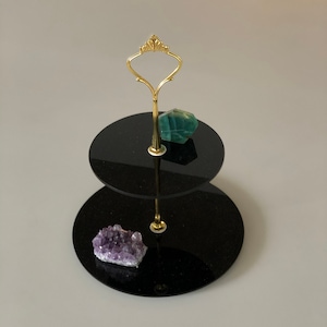 May include: Two-tier black acrylic cake stand with a gold metal handle. The top tier has a green crystal and the bottom tier has a purple crystal.