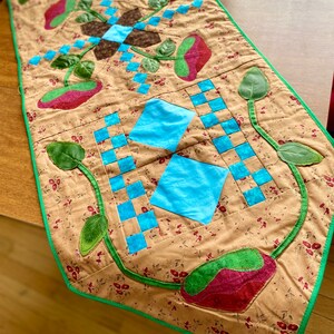 May include: A brown quilted table runner with a green border. The runner features a repeating pattern of blue squares, green leaves, and red flowers.