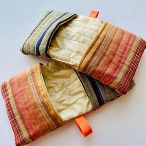 以下が含まれることがあります： ピンク、オレンジ、茶色、青の色合いのストライプ模様の、2つの長方形のキルト生地ポーチ。各ポーチはクリーム色の内側とオレンジ色のリボンループが付いています。