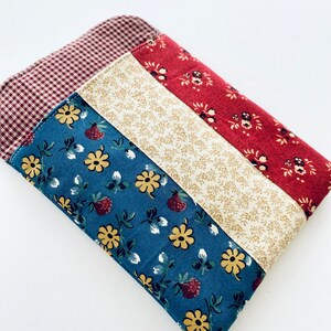 May include: A quilted fabric item with a patchwork design. The top left corner features a red and white gingham pattern. The design includes stripes of blue floral, cream floral, and red floral fabrics. The item is likely a small pouch or case.