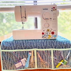 May include: A white Brother sewing machine with a floral design on the side, sitting on a blue and white patterned fabric sewing machine table with clear pockets for sewing supplies. The machine has a dial with stitch options and the model number is "JX2517".