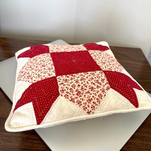 May include: Square decorative pillow with a patchwork design. The pillow features a central red square with white polka dots, surrounded by red floral print and cream-colored fabric. The edges are finished with a cream-colored border.