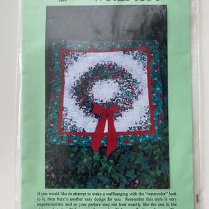 May include: A square quilt pattern titled CHRISTMAS WREATH featuring a central floral wreath design on a white background with a red border and a red bow. The packaging includes text describing a wallhanging that measures approximately 32 inches square.
