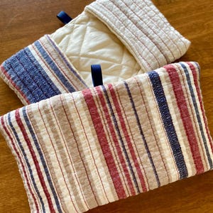 以下が含まれることがあります： 2つのキルト加工されたオーブンミトン。1つはクリーム色で赤いステッチと青いリボンループが付いています。もう1つは赤、青、ベージュのストライプ模様です。ミトンはキッチンでの使用を目的としています。