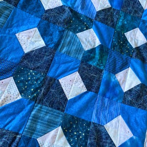May include: A handmade quilt featuring a patchwork design in various shades of blue. The quilt incorporates geometric shapes, including diamonds and squares, with a mix of patterned and solid fabrics. The color palette includes light, medium, and dark blues.