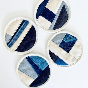 Puede incluir: Cuatro posavasos redondos de mezclilla con borde blanco. Cada posavasos está hecho de diferentes tonos de tela de mezclilla azul cosidos juntos en un patrón geométrico.