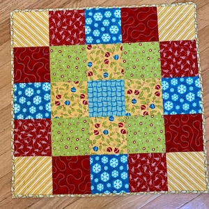 May include: A square, patchwork Christmas-themed table runner. The design features alternating squares of red, green, blue, and yellow fabrics with festive patterns like snowflakes, ornaments, and stripes. The edges are finished with a patterned border.