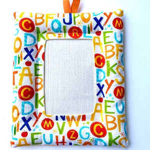 May include: A rectangular fabric picture frame featuring a white background with a colorful alphabet print including red, blue, green, and orange letters, numbers, and small icons. An orange ribbon loop is attached to the top center for hanging.
