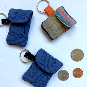 Puede incluir: Tres pequeñas bolsas de tela con llaveros. Las bolsas son azules, azules con ribete negro y beige con ribete naranja. Las bolsas están acolchadas y tienen un cierre de solapa. Las bolsas están sobre una superficie blanca.