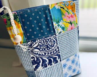 Op de afbeelding: Een patchwork crossbody tas met verschillende blauwe en bedrukte stoffen. De tas heeft bloemen-, paisley- en vichy-patronen. Een crèmekleurig kralenaccent hangt aan de riem.