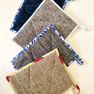 May include: Four square quilted pot holders in shades of blue, gray, and brown. Each pot holder has a different fabric pattern and a contrasting patterned border. A loop is attached to one corner for hanging.