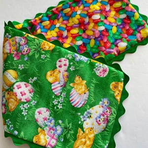 Può includere: Due quadrati di tessuto a tema pasquale. Uno presenta un motivo di colorate caramelle gommose. L'altro ha uno sfondo verde con uova di Pasqua, pulcini e dettagli floreali. Entrambi hanno un bordo verde smerlato.