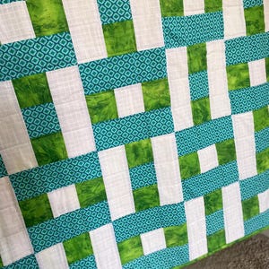 May include: A handmade quilt with a geometric pattern. The design features white, green, and teal blocks arranged in a grid. The green blocks have a mottled texture, while the teal sections have a diamond pattern. The quilt is likely made of cotton.