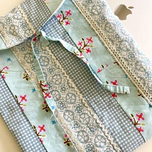 Puede incluir: Una funda para portátil de patchwork con un diseño floral y de cuadros vichy en azul claro. La funda está adornada con un ribete de encaje blanco y un cierre de lazo a juego. Flores rosas y hojas verdes están impresas en la tela azul claro.