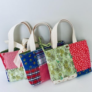 Puede incluir: Tres bolsos tote patchwork con asas color crema. Las bolsas presentan varios patrones de tela, incluyendo cuadros rojos, florales y cuadros escoceses. Una bolsa tiene una cinta rosa atada al asa. Las bolsas están dispuestas una al lado de la otra.