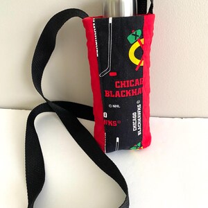 May include: A quilted fabric water bottle carrier featuring a black center panel with Chicago Blackhawks team graphics, including hockey sticks and text, flanked by solid red sides. It includes a black adjustable strap and a silver metal bottle.