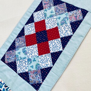 May include: A blue and white table runner with a red and blue patchwork design. The runner is made of fabric squares sewn together in a diamond pattern.