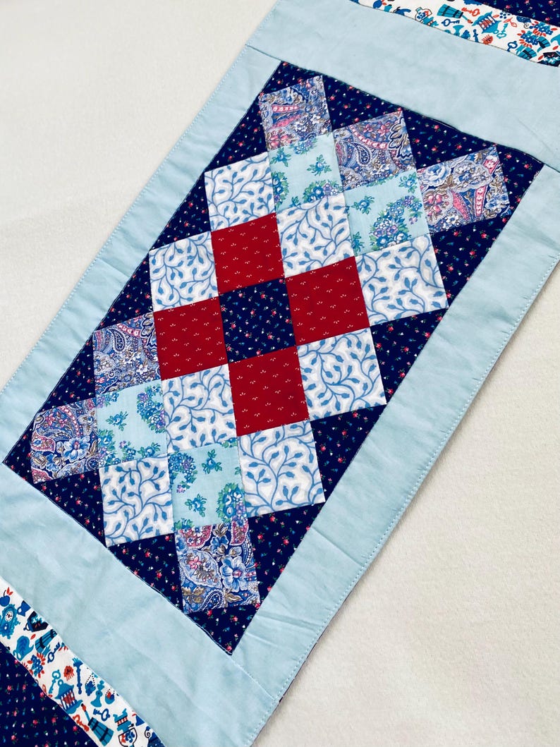 Pode incluir: Um corredor de mesa azul e branco com um design de patchwork vermelho e azul. O corredor &eacute; feito de quadrados de tecido costurados juntos em um padr&atilde;o de diamante.