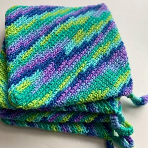 Könnte beinhalten: Ein Stapel von drei handgefertigten Topflappen aus Häkelarbeit in Blau-, Grün- und Lilatönen. Die Topflappen haben ein diagonales Streifenmuster und eine Schlaufe zum Aufhängen. Die Häkelarbeit ist engmaschig und die Farben sind lebendig.