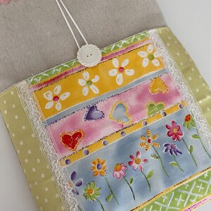 May include: A fabric pouch featuring a beige linen top section with pink rickrack trim. The lower section displays horizontal panels of yellow, pink, and blue fabric with floral and heart prints, accented by white lace trim and a white button closure.