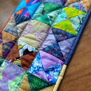 May include: A handmade patchwork quilt featuring a vibrant array of colors and patterns. The quilt is composed of numerous triangular fabric pieces in shades of blue, green, purple, and gold, creating a geometric design. The edges are finished with a blue polka dot fabric.