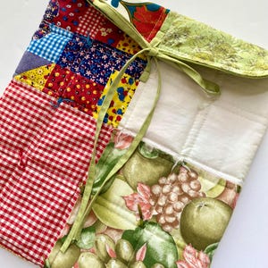 Puede incluir: Una funda de tela acolchada con un diseño de patchwork, con cuadros rojos, estampados florales y de frutas. La funda está atada con una cinta verde y tiene un interior acolchado blanco.