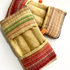 May include: Two quilted oven mitts in the image. The mitts are primarily beige with striped accents in red, green, and orange. Each mitt has a small orange loop for hanging. The fabric appears to be a textured cotton or linen.