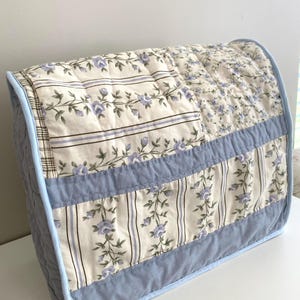 Peut inclure: Une housse d'appareil matelassée présentant un motif patchwork avec des fleurs crème, bleu clair et lavande. La housse comprend des rayures verticales, de petites fleurs bleues et des panneaux en tissu bleu clair uni avec un passepoil bleu.