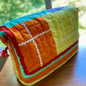 May include: A brightly coloured, quilted fabric item in a rectangular shape. It has a patchwork design with vibrant orange, yellow, turquoise, and red hues. White zigzag ribbon accents the orange sections. A small red tassel hangs from one end.