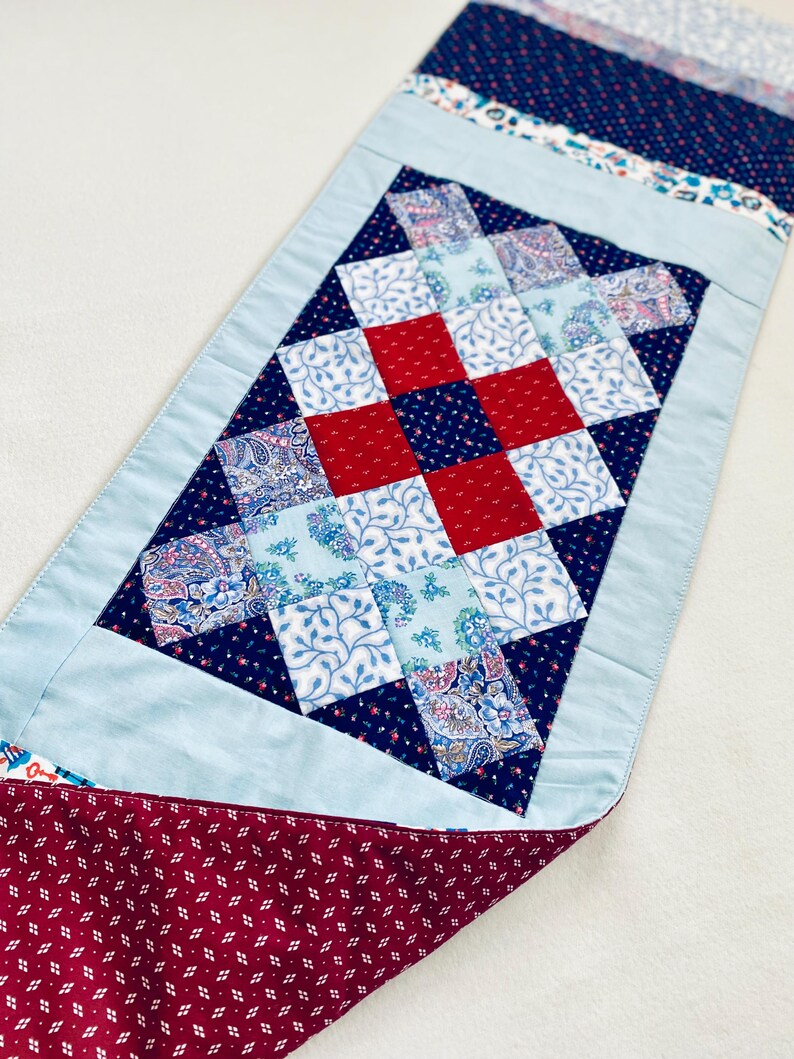 Pode incluir: Um corredor de mesa de patchwork azul e vermelho com um padr&atilde;o de diamante. O corredor tem uma borda azul claro e um verso bord&ocirc;.