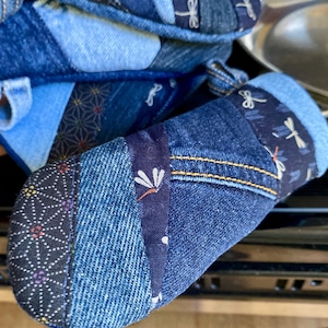 Puede incluir: Primer plano de dos manoplas de cocina de mezclilla. Las manoplas están hechas de mezclilla patchwork en varios tonos de azul, con costuras decorativas y detalles de tela de libélulas. Las manoplas están diseñadas para uso en la cocina.