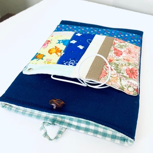 Peut inclure: Une pochette en tissu bleu avec une bordure à carreaux vert et blanc, un bouton marron et une anse en boucle. La pochette est ouverte et contient une pochette en tissu blanc à motif floral, une pochette en tissu blanc à motif bleu et blanc et une pochette en tissu blanc à motif jaune et bleu.