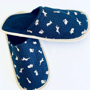 Pu&ograve; includere: Un paio di pantofole blu con un motivo di coniglio bianco. Le pantofole hanno un bordo color crema.
