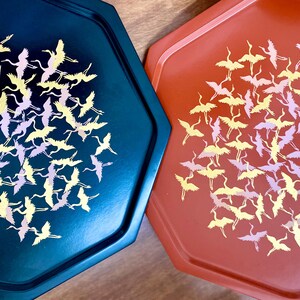 May include: Two hexagonal trays, one teal and one coral, are displayed. Each tray features a flock of stylized cranes in yellow and lavender. The trays have a glossy finish and are likely made of metal or a similar material.
